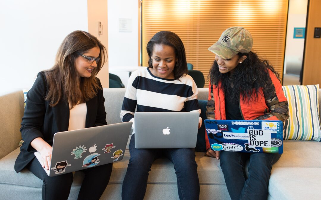 Photo shows three women of color sitting on a couch side by side with laptops.