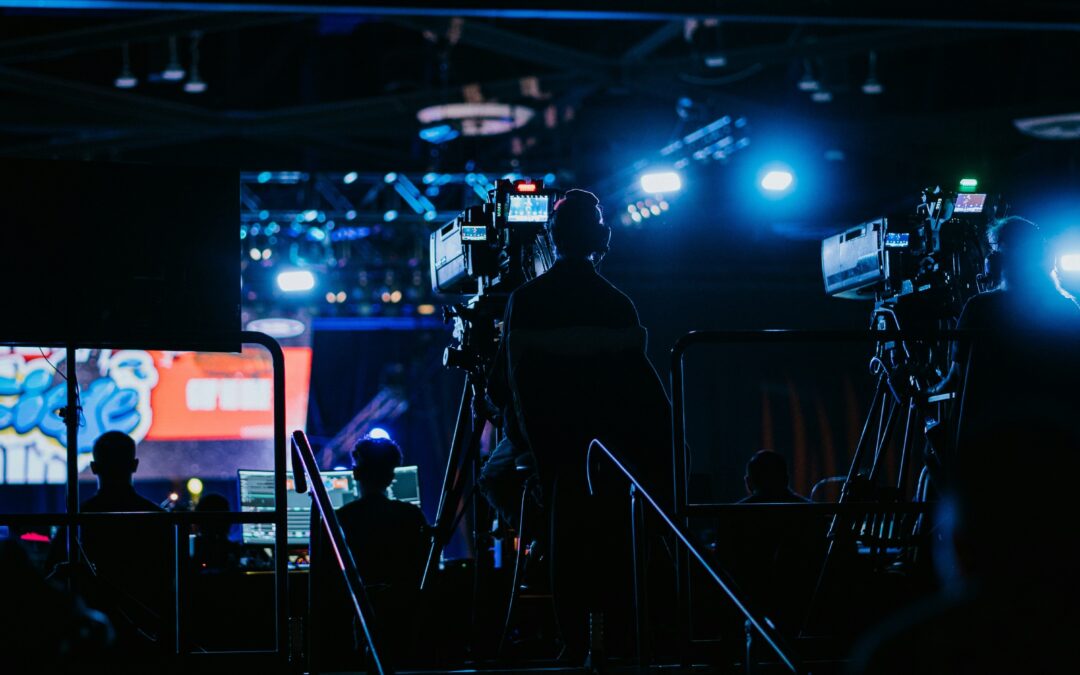 Silhouettes of camera operators and crew filming on a stage with bright blue lights and large screens in the background.